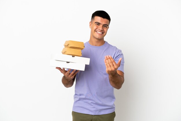 Jovem bonito segurando pizzas e hambúrgueres sobre fundo branco isolado, convidando para vir com a mão. Feliz que você veio