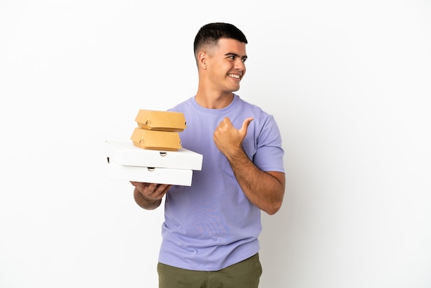 Jovem bonito segurando pizzas e hambúrgueres sobre um fundo branco isolado, apontando para o lado para apresentar um produto