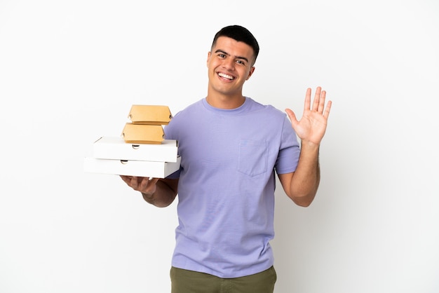 Jovem bonito segurando pizzas e hambúrgueres sobre um fundo branco isolado e saudando com a mão com uma expressão feliz