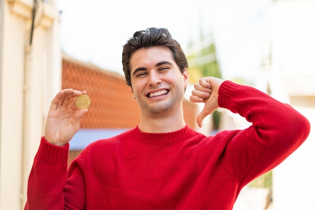 Jovem bonito segurando um Bitcoin ao ar livre orgulhoso e satisfeito