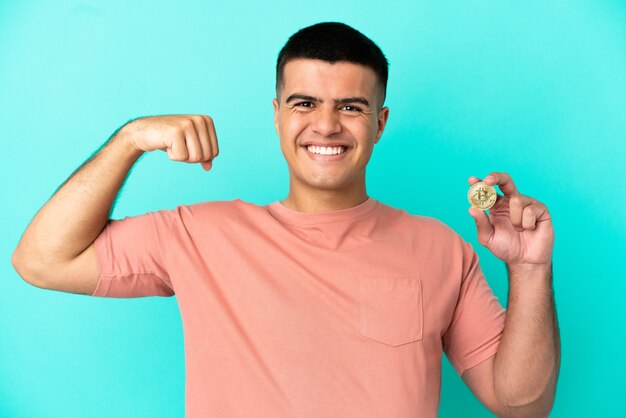 Jovem bonito segurando um Bitcoin sobre um fundo azul isolado fazendo um gesto forte