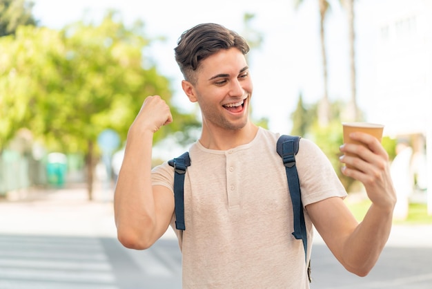 Jovem bonito segurando um café para viagem ao ar livre comemorando uma vitória