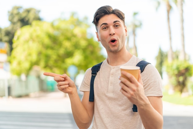 Jovem bonito segurando um café para viagem ao ar livre surpreso e apontando para o lado