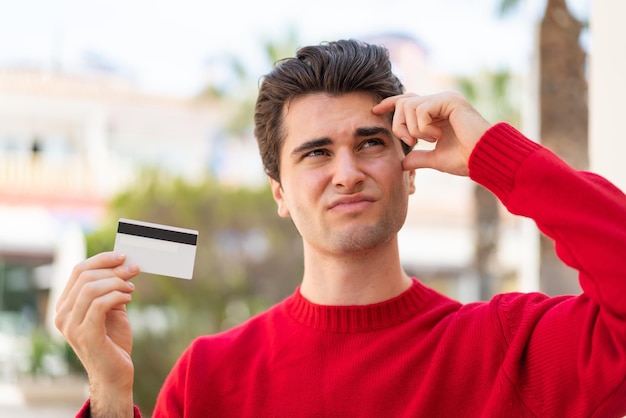 Foto jovem bonito segurando um cartão de crédito ao ar livre com dúvidas e com expressão facial confusa