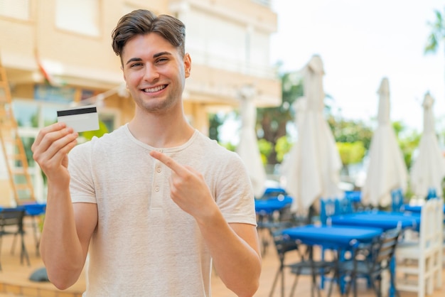 Jovem bonito segurando um cartão de crédito ao ar livre e apontando-o