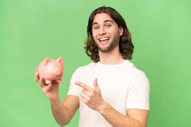 Jovem bonito segurando um cofrinho sobre fundo isolado e apontando-o