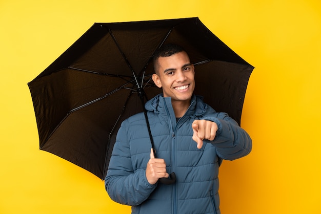 Jovem bonito segurando um guarda-chuva sobre parede amarela isolada aponta o dedo para você com uma expressão confiante