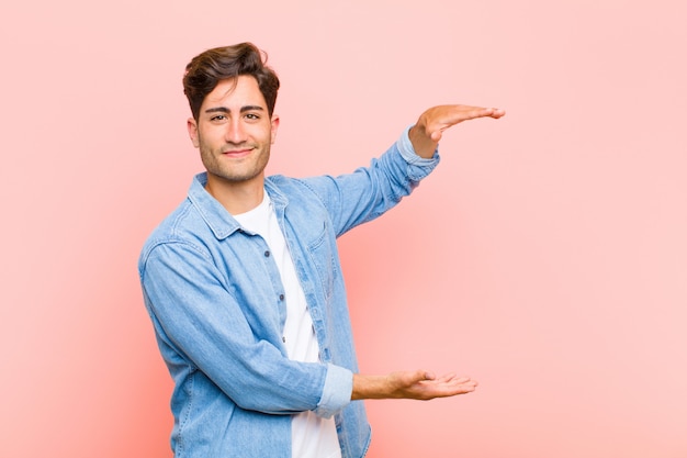 Jovem bonito segurando um objeto com as duas mãos no espaço da cópia lateral, mostrando, oferecendo ou anunciando um objeto na rosa