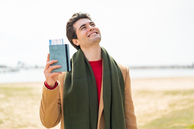 Jovem bonito segurando um passaporte ao ar livre olhando para cima enquanto sorri