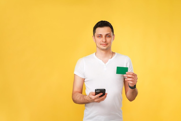 Foto jovem bonito segurando um telefone e um cartão bancário na mão em uma camiseta branca sobre um fundo amarelo