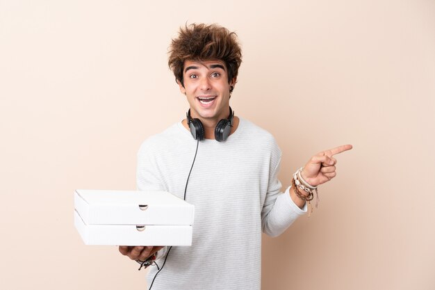 Jovem bonito segurando uma pizza sobre parede isolada surpreendeu e apontando o dedo para o lado