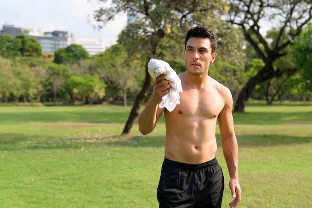 Jovem bonito sem camisa hispânico segurando uma camisa no parque