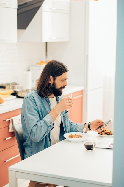 Jovem bonito sentado à mesa com um copo de suco e olhando para a tela de um smartphone moderno