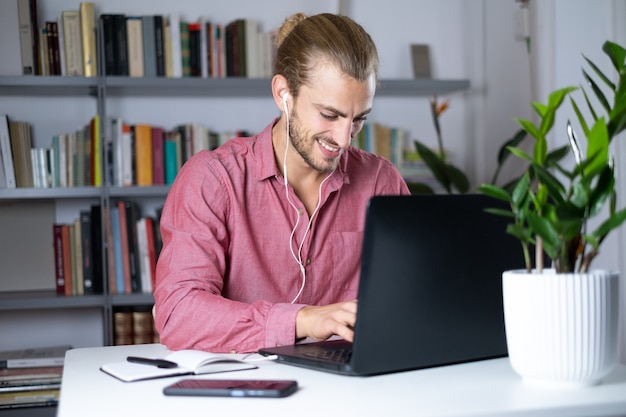 Jovem bonito sentado à mesa em casa trabalhando com um laptop e se sentindo feliz