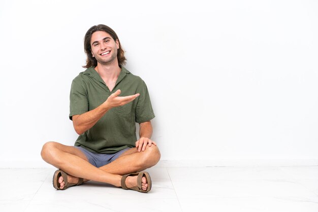 Jovem bonito sentado no chão isolado no fundo branco apresentando uma ideia enquanto olha sorrindo para