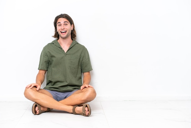 Jovem bonito sentado no chão isolado no fundo branco com expressão facial de surpresa