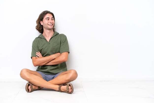 Jovem bonito sentado no chão isolado no fundo branco com os braços cruzados e feliz