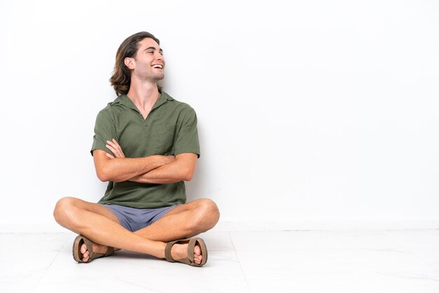 Jovem bonito sentado no chão isolado no fundo branco feliz e sorridente