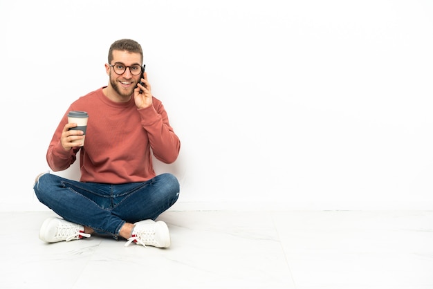 Jovem bonito sentado no chão segurando um café para levar e um celular