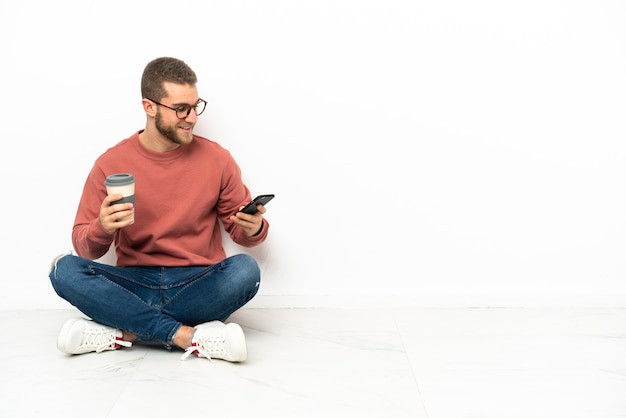 Jovem bonito sentado no chão segurando um café para levar e um celular