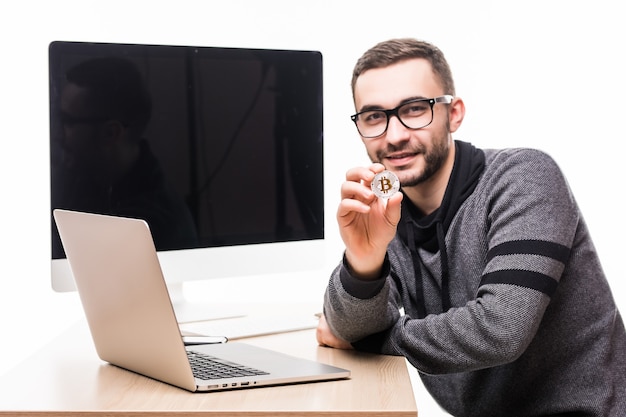 Jovem bonito sentado no escritório com o laptop e a tela do monitor nas costas apontando bitcoin em branco