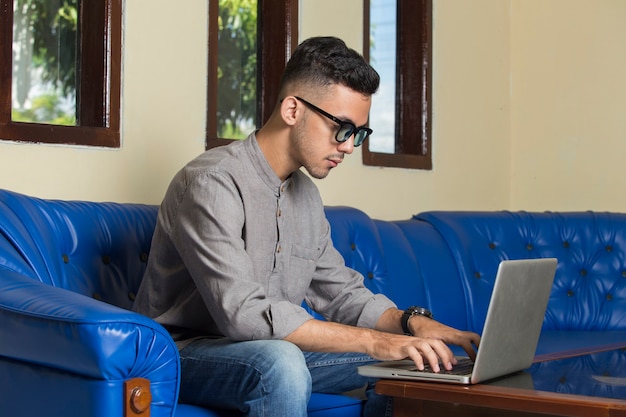 Jovem bonito sentado no sofá em casa trabalhando em um laptop online