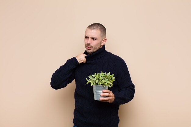 Jovem bonito, sentindo-se estressado, ansioso, cansado e frustrado, puxando o pescoço da camisa, olhando frustrado com o problema segurando uma planta