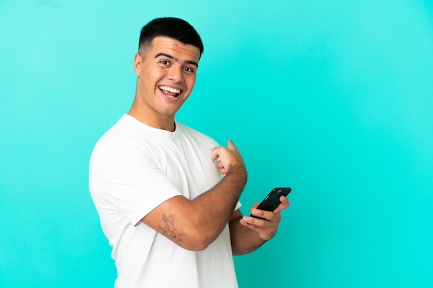 Jovem bonito sobre fundo azul isolado usando telefone celular e apontando para trás