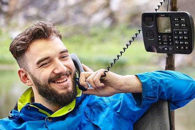 Jovem bonito sorri durante uma agradável conversa telefônica na natureza, usando um telefone com botão de pressão desatualizado com fio trançado.