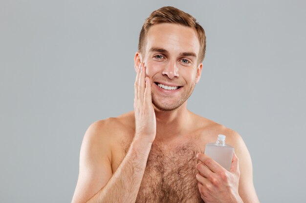 Jovem bonito sorridente aplicando loção após a barba no rosto sobre uma parede cinza