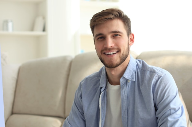 Foto jovem bonito sorridente homem sentado em um sofá em sua sala de estar e olhando para a câmera.