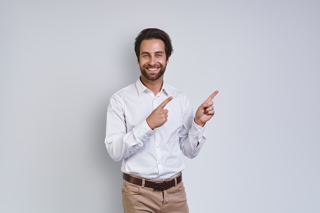 Jovem bonito sorridente na camisa branca, olhando para a câmera e apontando para longe em pé contra ...