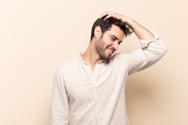 Jovem bonito sorrindo alegre e casualmente, levando a mão na cabeça com um olhar positivo, feliz e confiante