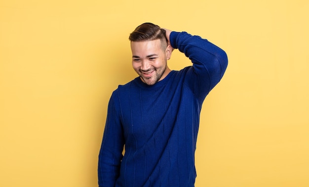 Jovem bonito sorrindo alegre e casualmente, levando a mão na cabeça com um olhar positivo, feliz e confiante