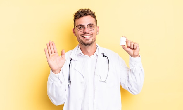 Jovem bonito sorrindo alegremente, acenando com a mão, dando as boas-vindas e cumprimentando você. conceito de frasco de comprimidos para médico