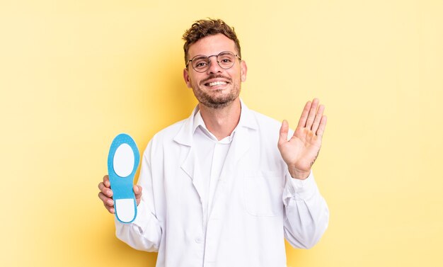 Jovem bonito sorrindo alegremente, acenando com a mão, dando as boas-vindas e cumprimentando você. conceito de quiropodista