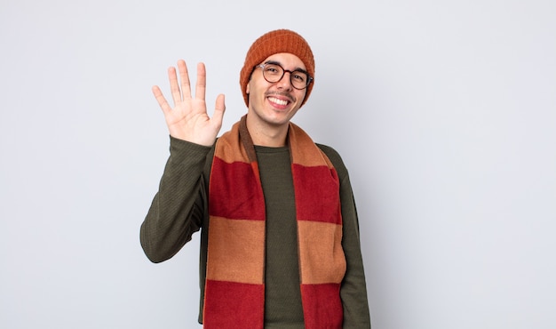 Jovem bonito sorrindo alegremente, acenando com a mão, dando as boas-vindas e cumprimentando você. conceito de roupas de inverno