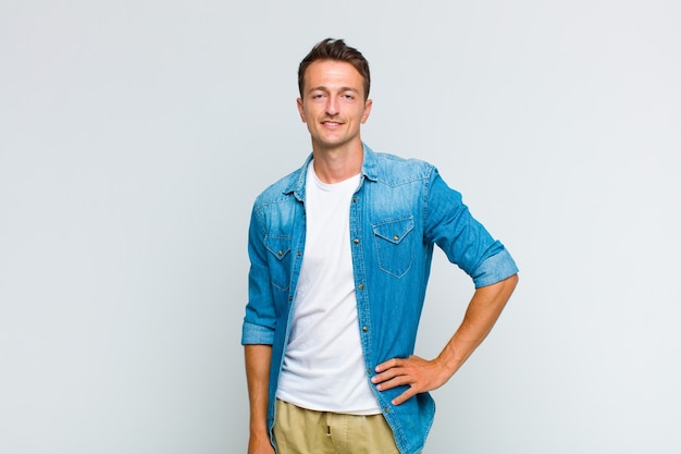 Jovem bonito sorrindo alegremente com uma mão no quadril e atitude confiante, positiva, orgulhosa e amigável