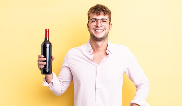 Jovem bonito sorrindo alegremente com uma mão no quadril e confiante. conceito de garrafa de vinho