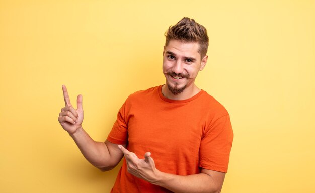 Jovem bonito sorrindo alegremente e apontando para o lado e para cima com as duas mãos, mostrando o objeto no conceito de bigode estranho de espaço de cópia