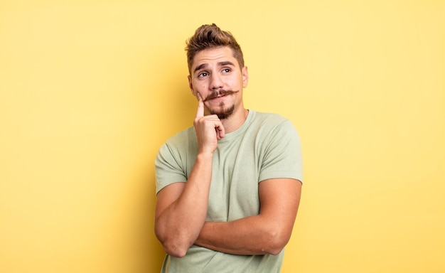 Jovem bonito sorrindo alegremente e sonhando acordado ou duvidando, olhando para o lado. conceito de bigode estranho