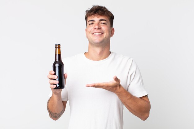 Jovem bonito sorrindo alegremente, se sentindo feliz e mostrando um conceito e segurando uma garrafa de cerveja