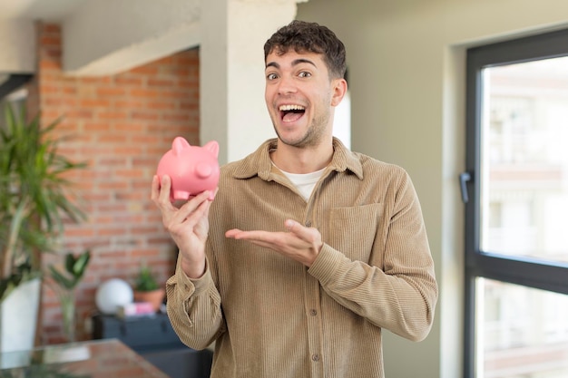 jovem bonito sorrindo alegremente, sentindo-se feliz e mostrando um conceito de cofrinho