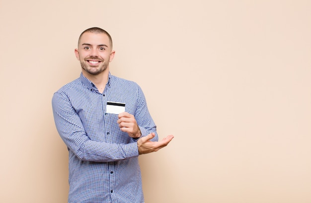 Jovem bonito sorrindo alegremente, sentindo-se feliz e mostrando um conceito no espaço da cópia com a palma da mão com cartão de crédito