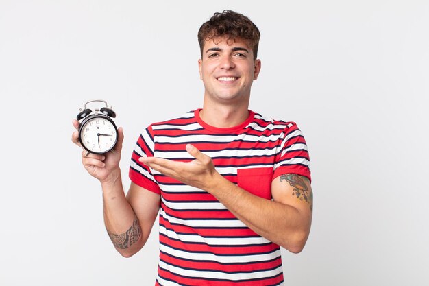 Jovem bonito sorrindo alegremente, sentindo-se feliz, mostrando um conceito e segurando um despertador