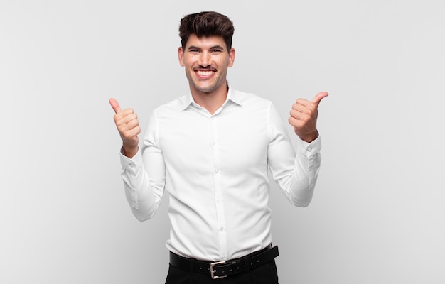 Jovem bonito sorrindo com alegria e parecendo feliz, sentindo-se despreocupado e positivo com os dois polegares para cima