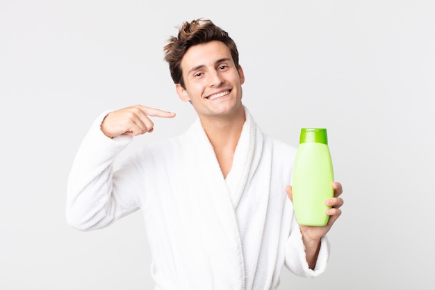 Jovem bonito sorrindo com confiança, apontando para o seu largo sorriso com um roupão de banho e segurando um frasco de xampu