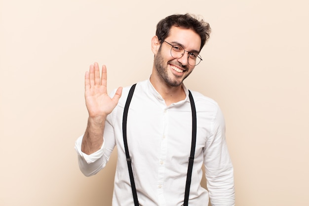 Jovem bonito sorrindo com felicidade e alegria, acenando com a mão, dando as boas-vindas e cumprimentando você ou dizendo adeus