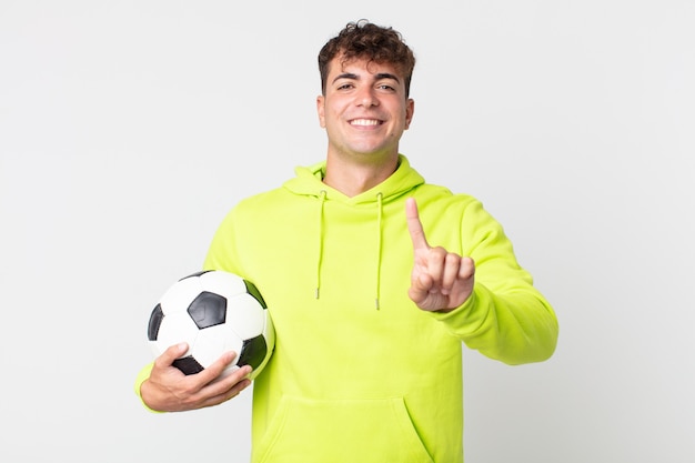 Jovem bonito sorrindo com orgulho e confiança, alcançando o primeiro lugar e segurando uma bola de futebol