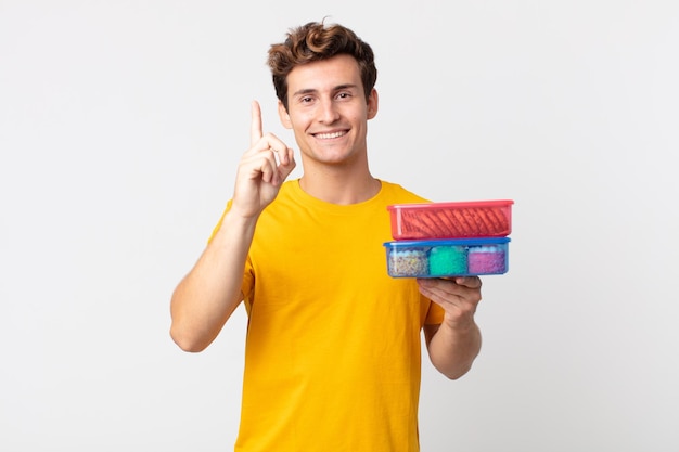 Jovem bonito sorrindo com orgulho e confiança fazendo o número um e segurando lancheiras